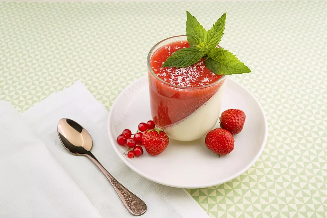 Ein Glas Panna Cotta mit Erdbeersauce, garniert mit Minzblättern und Kokosflocken, serviert mit Erdbeeren und roten Johannisbeeren auf einem weißen Teller – bringt einen Hauch Mallorca auf Ihren Tisch – neben einem Löffel und einer Serviette.