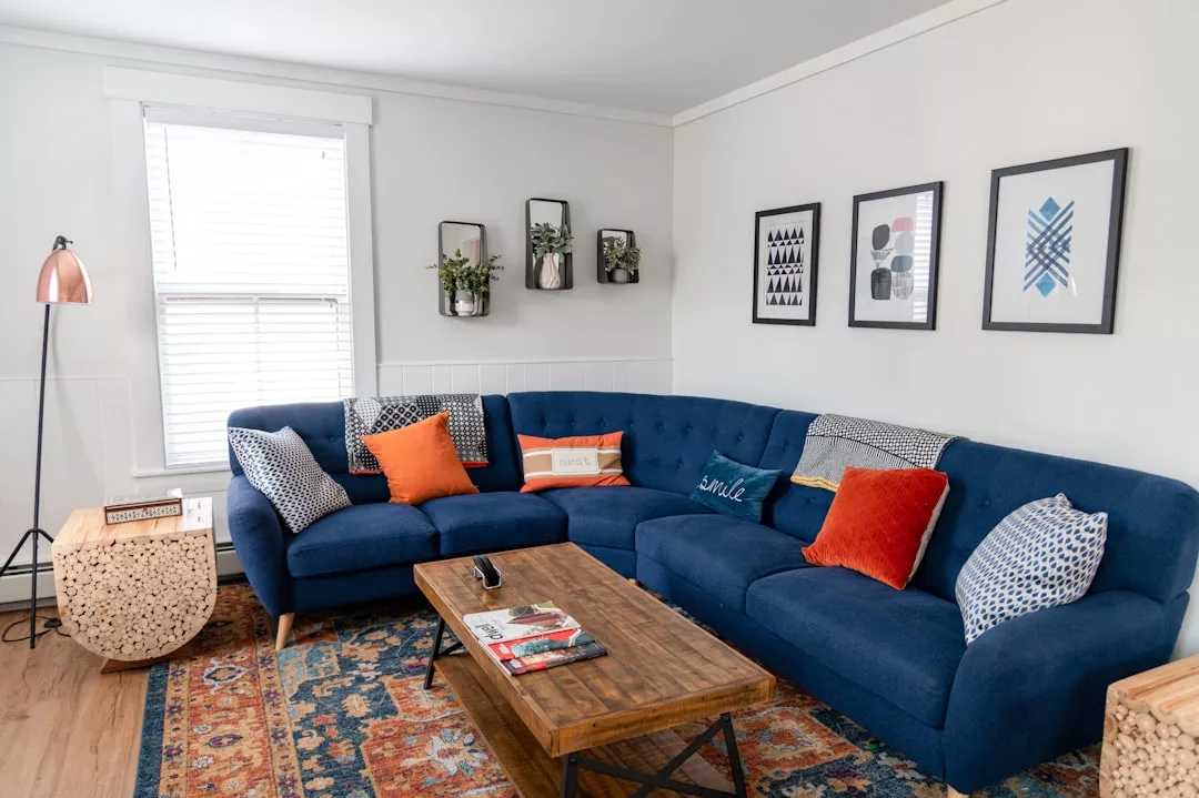Ein Wohnzimmer im Mallorca-Stil mit einem blauen Sofa, bunten Kissen, einem Couchtisch aus Holz, Wandkunst und Pflanzen an einer hellen Wand. Ein Fenster mit Jalousien und ein gemusterter Teppich runden das Küstenambiente ab.