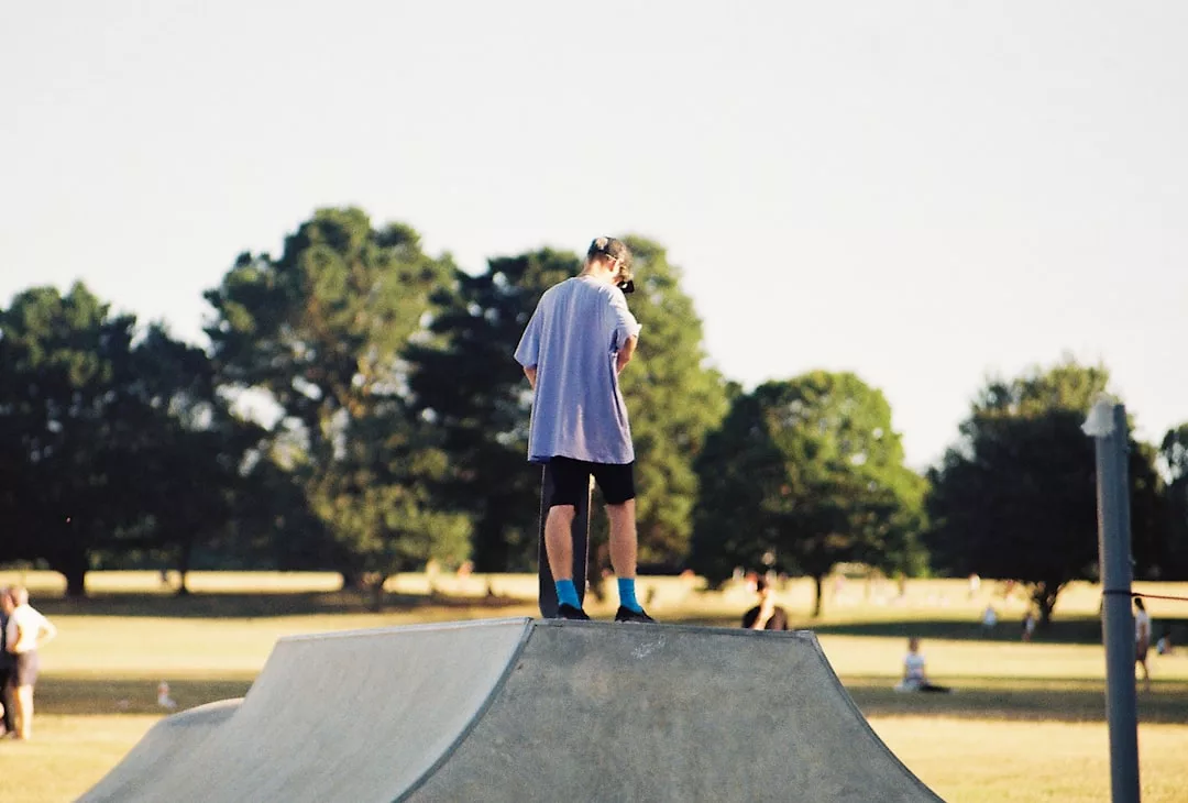 Eine Person in einem hellen Hemd und schwarzen Shorts steht auf einer Skaterampe aus Beton in einem Park auf Mallorca, umgeben von Bäumen und Menschen, die die lebhafte Atmosphäre genießen.