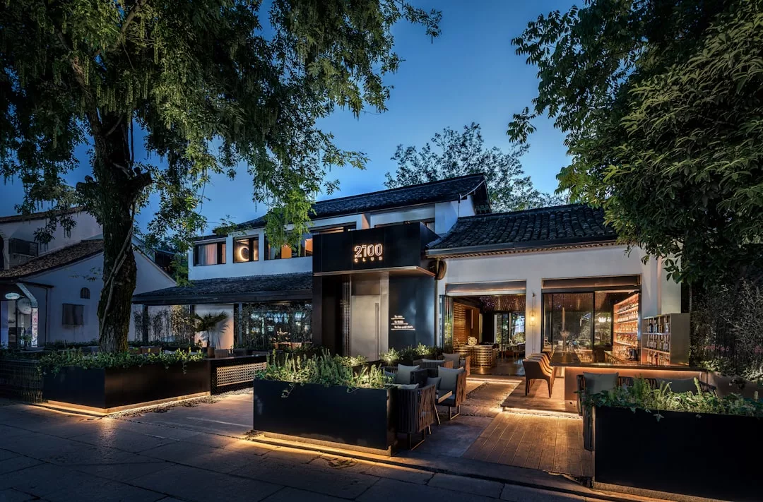 Außenansicht eines modernen Restaurants in der Abenddämmerung auf Mallorca mit Sitzgelegenheiten im Freien, großen Fenstern und warmer Beleuchtung, umgeben von Bäumen und üppigem Grün.