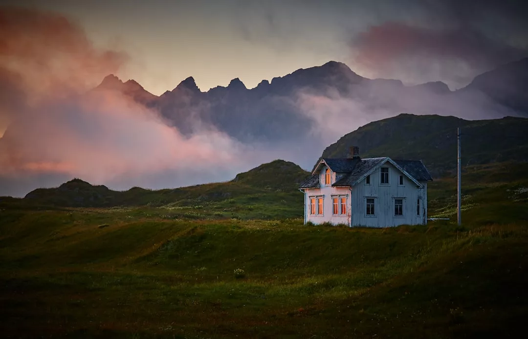 Ein weißes Haus mit erleuchteten Fenstern steht allein auf einer Wiese auf Mallorca, mit nebligen Bergen und einem farbenfrohen Himmel im Hintergrund.