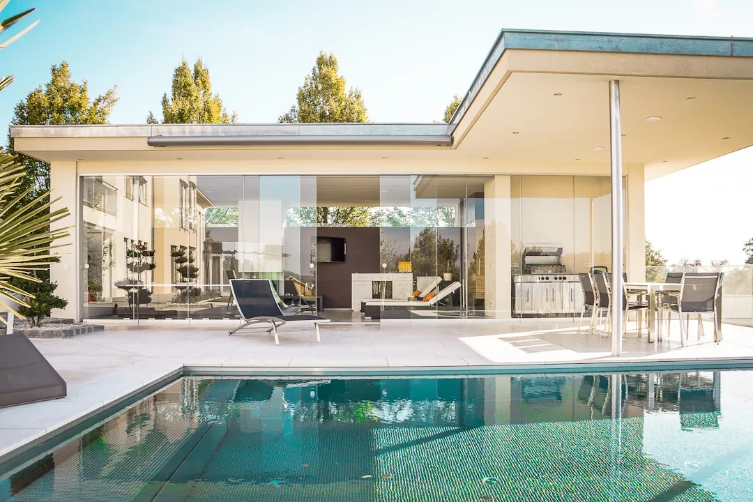Modernes Haus auf Mallorca mit großen Glasfenstern, Liegestühlen im Freien und einem Swimmingpool im Vordergrund unter klarem Himmel.