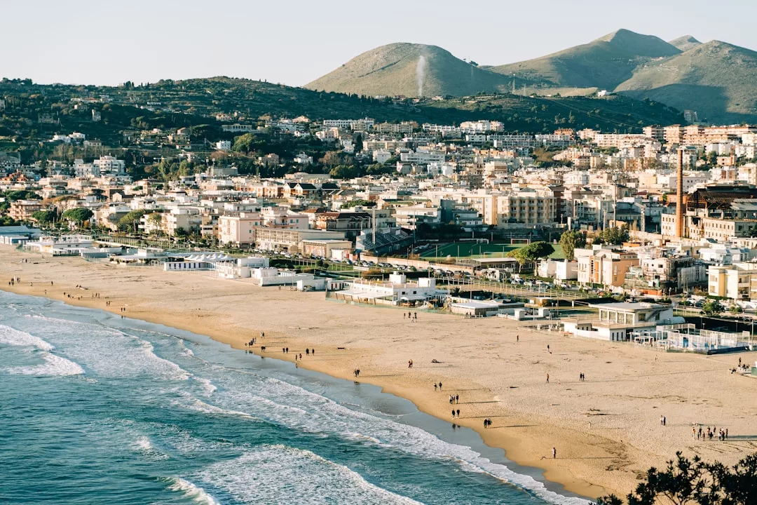 Eine Küstenstadt auf Mallorca mit Sandstränden, sanften Wellen und Menschen, die am Ufer entlang spazieren, eingerahmt von Gebäuden und grünen Hügeln im Hintergrund.