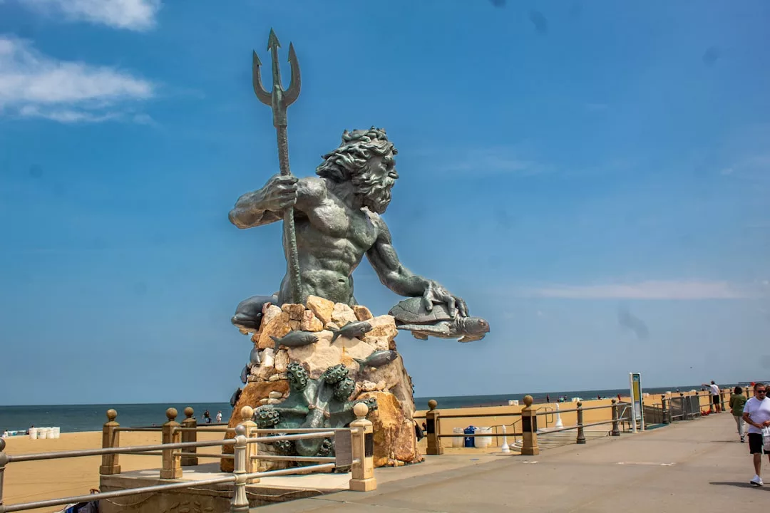Eine große Statue von Neptun mit einem Dreizack steht auf einem Steinsockel neben der Strandpromenade und dem Sandstrand unter einem blauen Himmel über Mallorca.