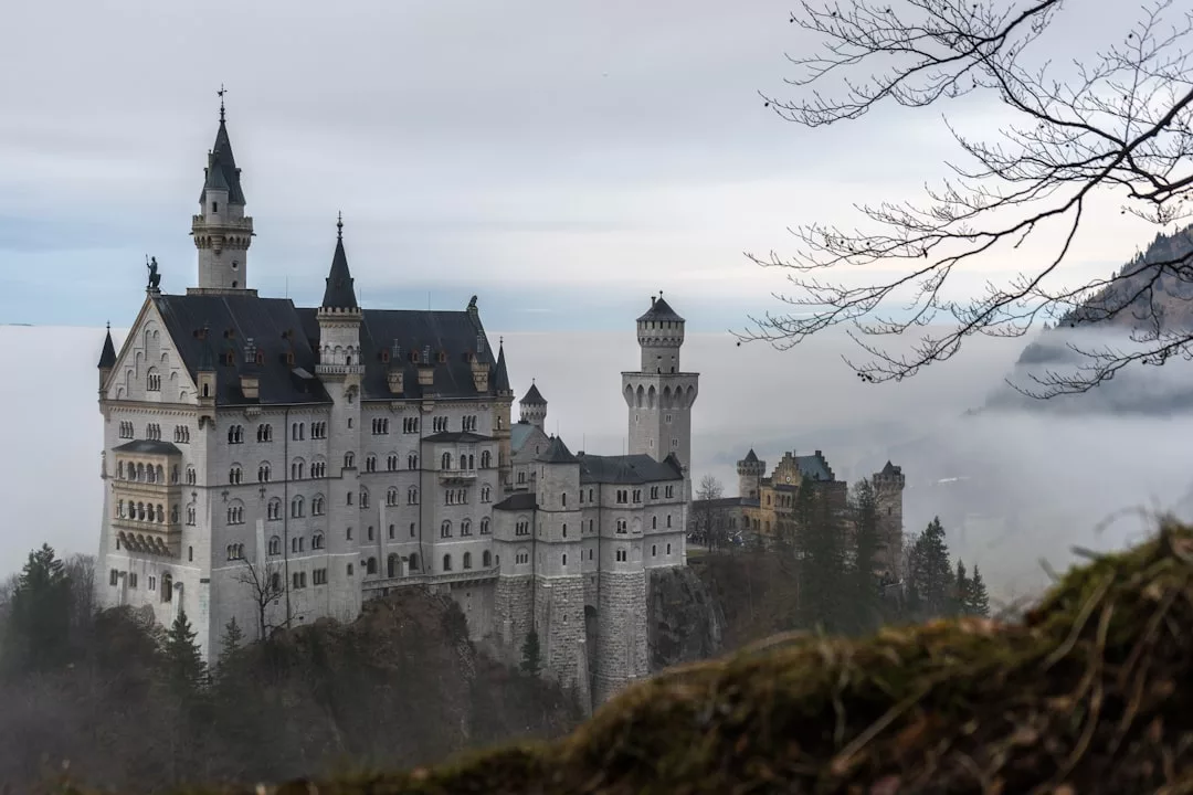 Auf einem Hügel auf Mallorca, umgeben von Nebel und Bäumen, unter einem bewölkten Himmel, steht eine große, reich verzierte Burg mit mehreren Türmen.