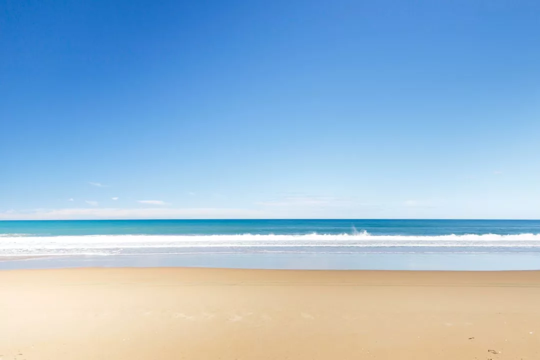 Ein klarer blauer Himmel über einem ruhigen Ozean mit sanften Wellen, die an einen leeren Sandstrand auf Mallorca schlagen.