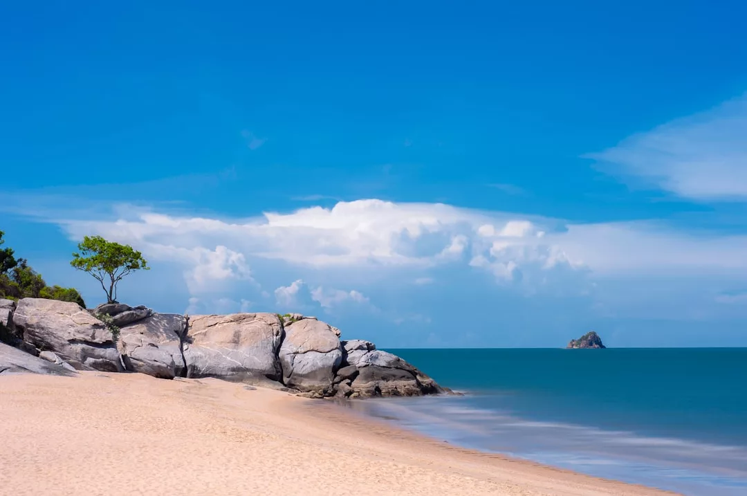 Ein Sandstrand auf Mallorca mit großen Felsen, ein paar kleinen Bäumen, ruhigem türkisfarbenem Wasser und einer kleinen Insel in der Ferne unter einem blauen Himmel mit vereinzelten Wolken.