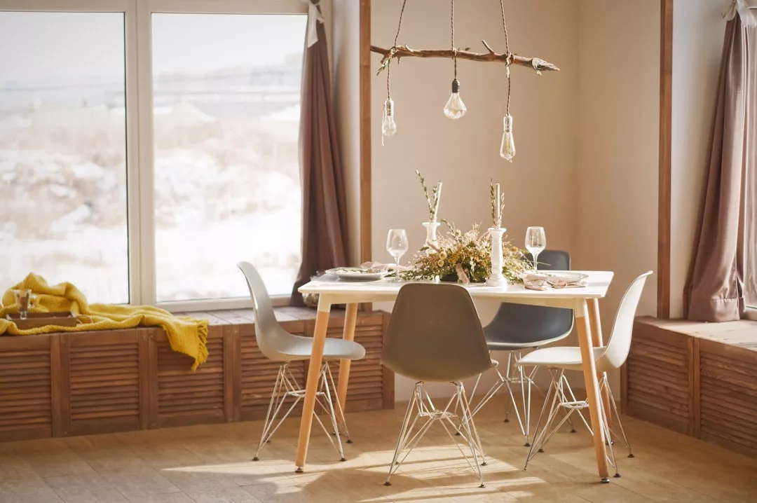 Darüber hängt ein Esstisch für vier Personen mit minimalistischen Stühlen, einem Blumengesteck, Weingläsern und Pendelleuchten; große Fenster lassen das natürliche Licht Mallorcas herein.