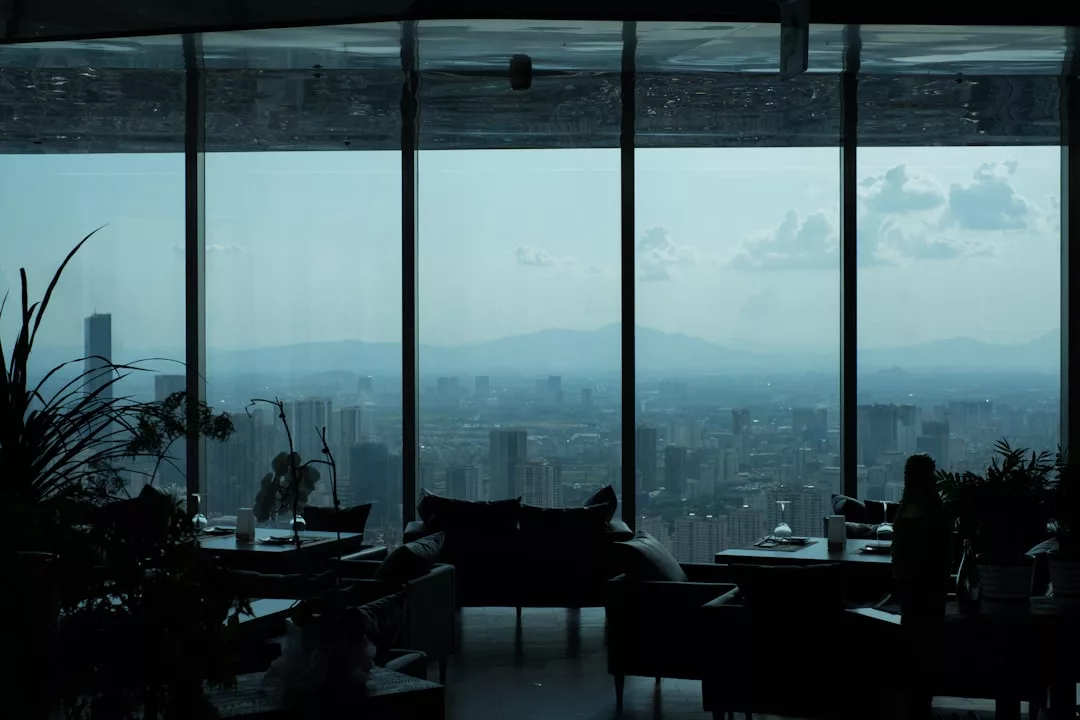 Ein Blick auf die Stadtlandschaft durch große, vom Boden bis zur Decke reichende Fenster in einem Hochhaus, mit Silhouetten von Zimmerpflanzen und Möbeln im Vordergrund, die an das moderne Leben auf Mallorca erinnern.