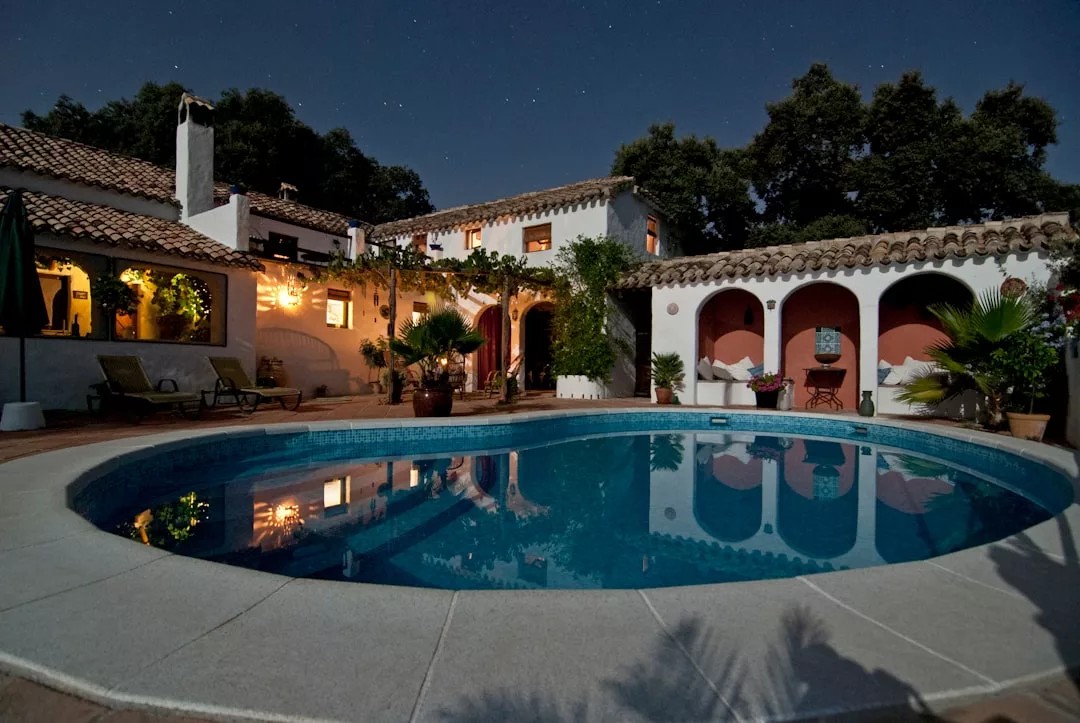 Eine Villa auf Mallorca mit Ziegeldach umgibt einen runden Pool, der nachts von Außenlichtern beleuchtet wird, mit Bäumen und einem Sternenhimmel im Hintergrund.