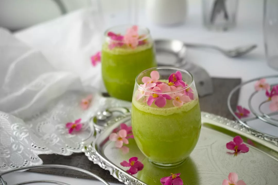 Zwei Gläser mit grünem Smoothie und essbaren rosa Blüten stehen auf einem silbernen Tablett und rufen den lebendigen Geist Mallorcas hervor. Weitere Blüten sind auf dem Tablett und dem Tisch verteilt.