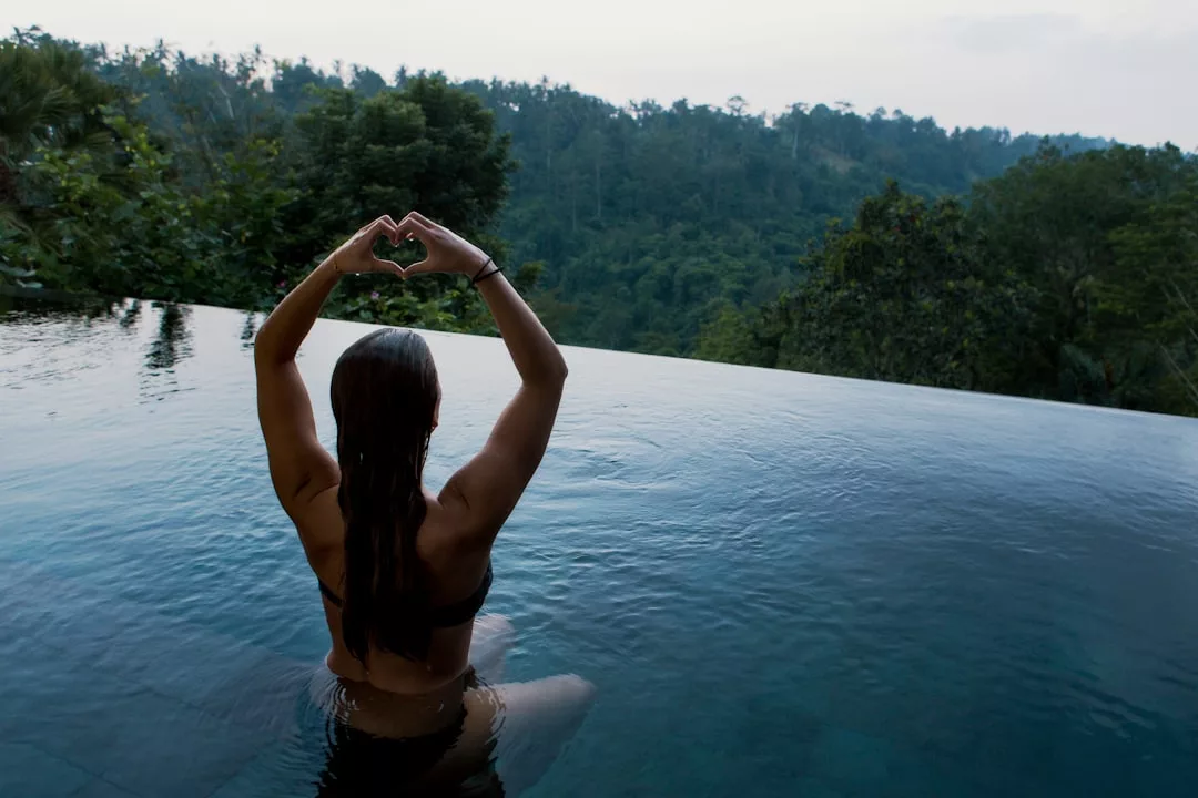 Eine Person steht in einem Infinity-Pool auf Mallorca, mit Blick auf üppige grüne Bäume, und hat die Arme so erhoben, dass sie mit ihren Händen die Form eines Herzens bilden.