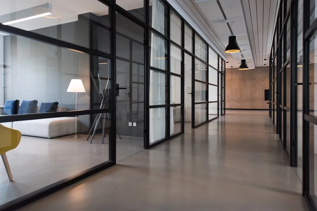 Ein moderner Büroflur im mallorquinischen Stil mit Glaswänden, einem grauen Sofa, einer weißen Stehlampe und minimalistischem Dekor. Über dem polierten Betonboden hängen schwarze Deckenleuchten.