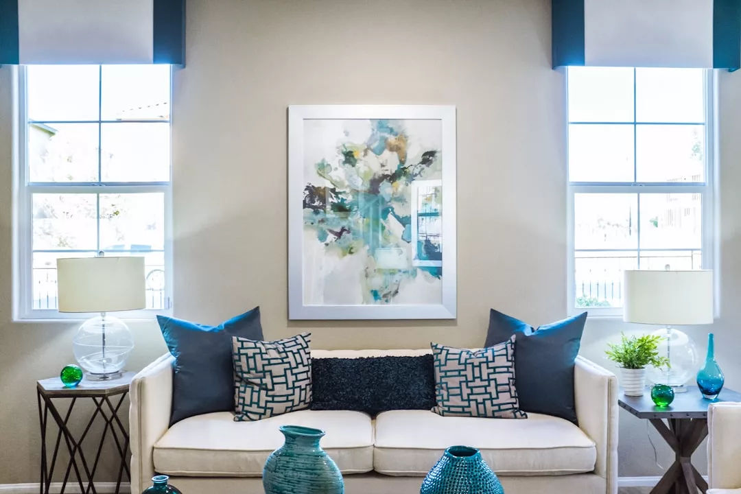 Ein modernes Wohnzimmer mit einem weißen Sofa, geometrischen blauen Kissen, Beistelltischen mit Lampen, dekorativen Vasen und abstrakter Kunst zwischen zwei großen Fenstern erinnert an die entspannte Eleganz Mallorcas.