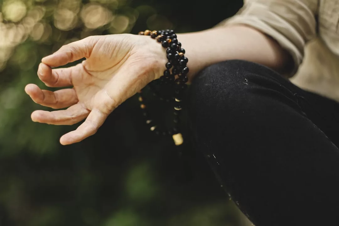 Eine Person sitzt im Schneidersitz, hält ihre Hand in einer Meditationsgeste und trägt mehrlagige schwarze Perlenarmbänder am Handgelenk, im Freien inmitten der üppigen Vegetation Mallorcas.