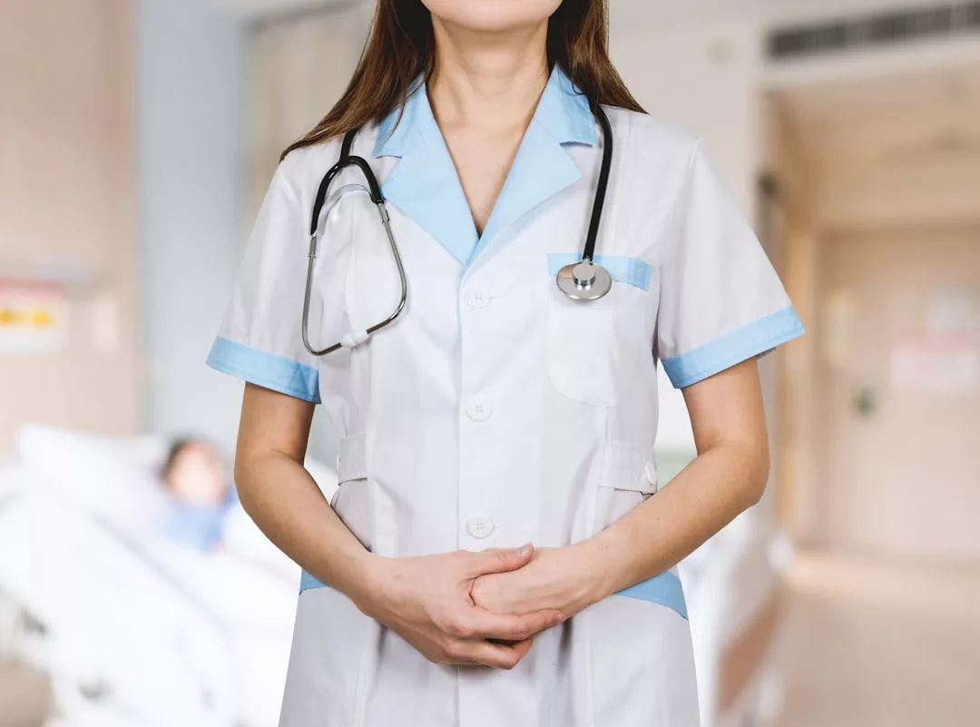 Ein Mediziner in weißer Uniform und mit einem Stethoskop steht mit gefalteten Händen in einem Krankenhauszimmer auf Mallorca; im unscharfen Hintergrund ist ein Patient zu sehen.