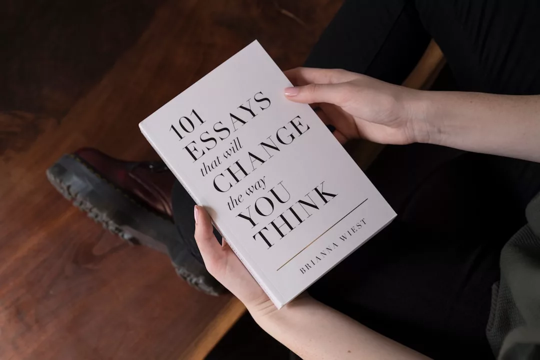 Eine Person hält das Buch „101 Essays That Will Change the Way You Think“ von Brianna Wiest in der Hand, während sie auf einem Holzboden sitzt und von Mallorca träumt.