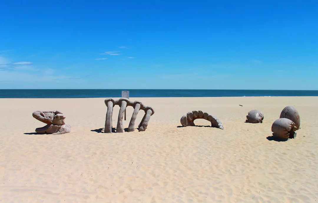 Eine große Skulptur einer teilweise vergrabenen Hand erhebt sich unter einem klaren blauen Himmel aus dem Sandstrand auf Mallorca, im Hintergrund ist das Meer zu sehen.