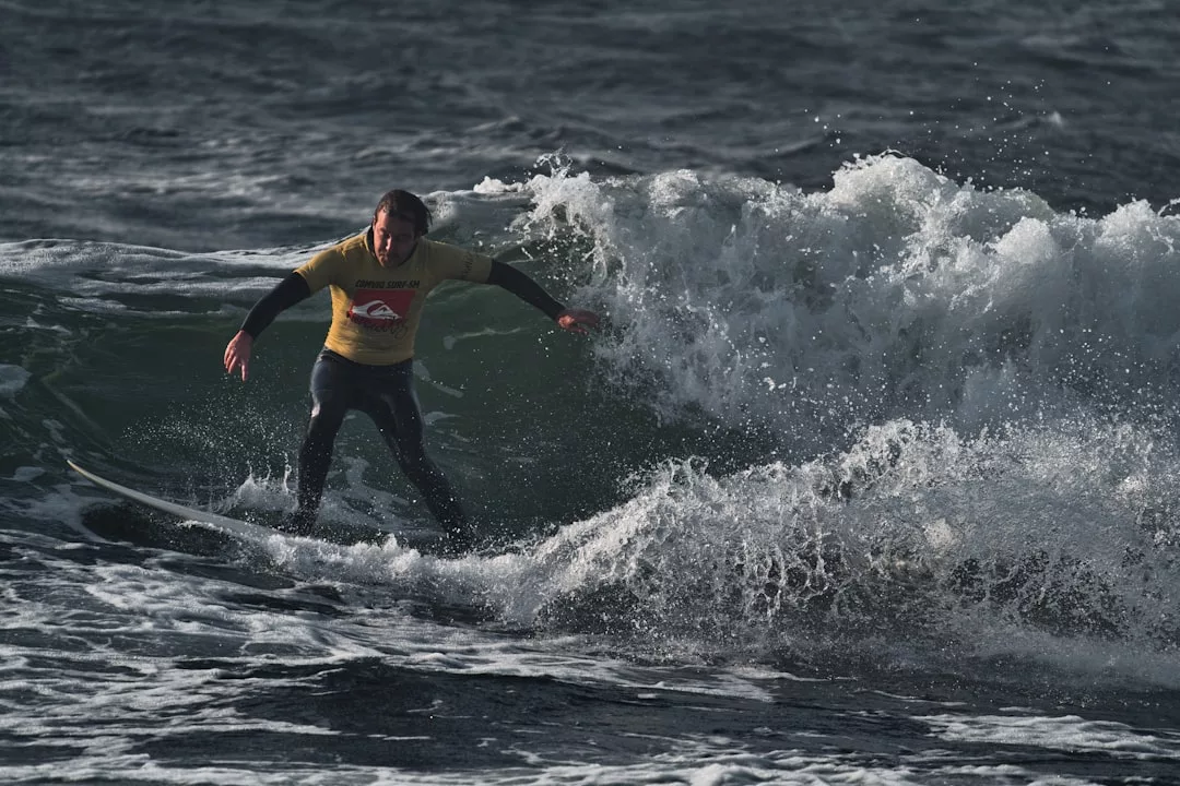 Ein Surfer in einem gelben Hemd und einem schwarzen Neoprenanzug reitet auf einer Welle in den rauen, dunklen Gewässern vor Mallorca.