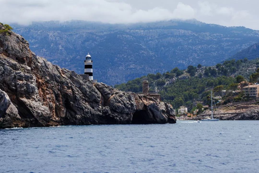 Ein schwarz-weiß gestreifter Leuchtturm steht auf felsigen Klippen am Meer auf Mallorca, mit Bergen und einigen Gebäuden im Hintergrund.