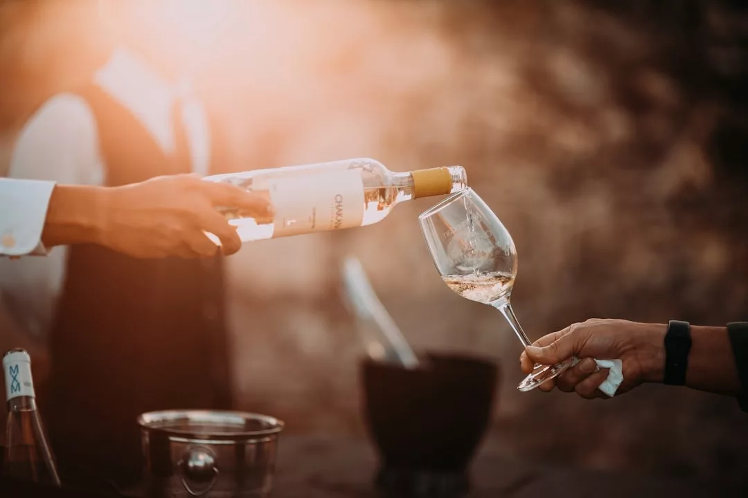 Eine Person gießt Weißwein in ein Glas, das eine andere Person hält. Ein unscharfer Hintergrund und sanftes Sonnenlicht fangen die entspannte Atmosphäre Mallorcas ein.