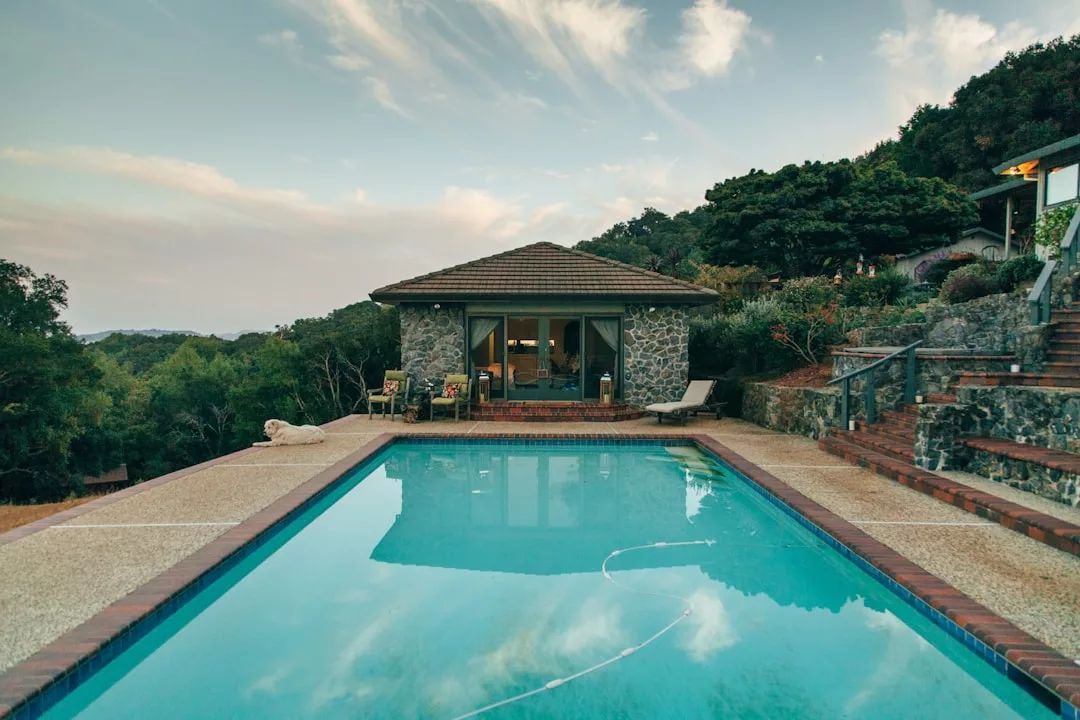 Ein rechteckiger Außenpool mit klarem Wasser, gesäumt von einem Steinweg, liegt vor einem Poolhaus aus Stein im mallorquinischen Stil, umgeben von Bäumen und einer hügeligen Landschaft.