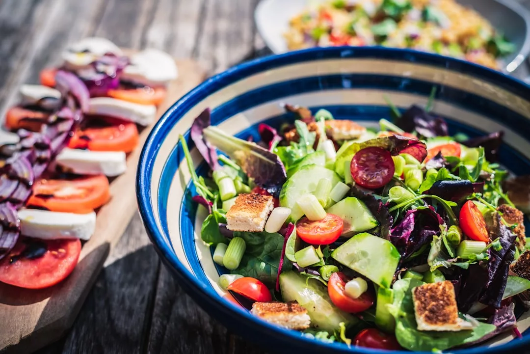 Eine Schüssel mit gemischtem Salat aus Tomaten, Gurken, Croutons und Gemüse steht auf einem Holztisch neben einer Platte mit geschnittenen Tomaten, Mozzarella und Basilikum – und fängt die frischen Aromen ein, die von Mallorca inspiriert sind.