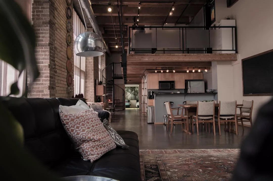 Modernes Loft-Apartment auf Mallorca mit unverputzten Ziegelwänden, hohen Decken, einem Zwischengeschoss, einer stilvollen Küche und einem Essbereich mit einem schwarzen Ledersofa und gemusterten Kissen im Vordergrund.