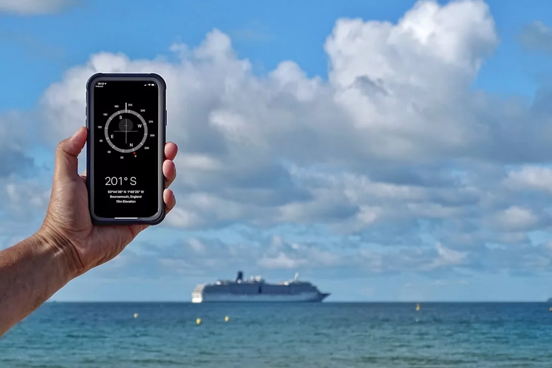Eine Hand hält ein Smartphone mit einer Kompass-App in Richtung Meer und zeigt 201° S in Richtung Mallorca; im Hintergrund fährt ein großes Kreuzfahrtschiff unter einem teilweise bewölkten Himmel.