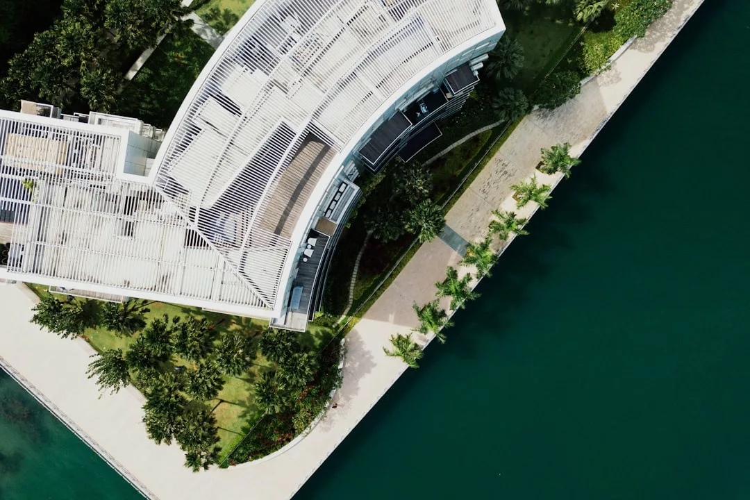 Luftaufnahme eines modernen Gebäudes mit weißem Dach auf Mallorca, neben einem angelegten Gehweg und grünem Wasser.
