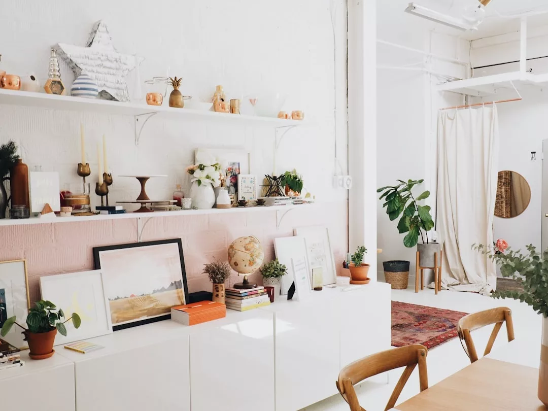 Ein heller, minimalistischer Raum im mallorquinischen Stil mit weißen Wänden, Holzstühlen, einem langen weißen Schrank, Zimmerpflanzen, Büchern, gerahmter Kunst und Deko-Objekten auf Regalen und Oberflächen.