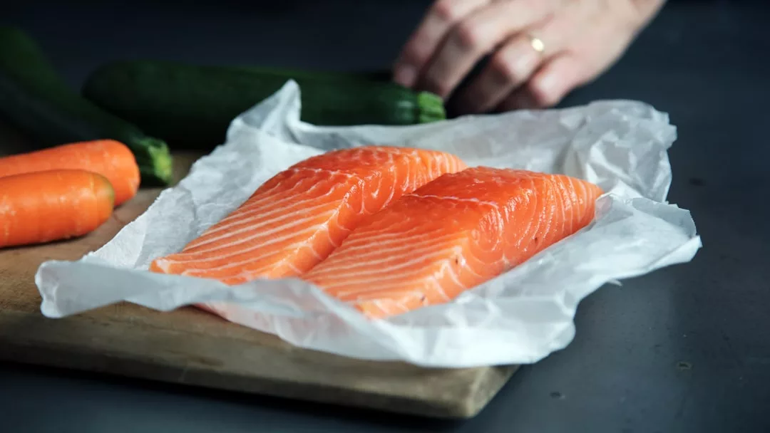 Zwei rohe Lachsfilets auf Pergamentpapier, auf einem Holzbrett platziert, mit Karotten, einer Zucchini und der Hand eines Menschen im Hintergrund – eine Erinnerung an die frischen Aromen Mallorcas.