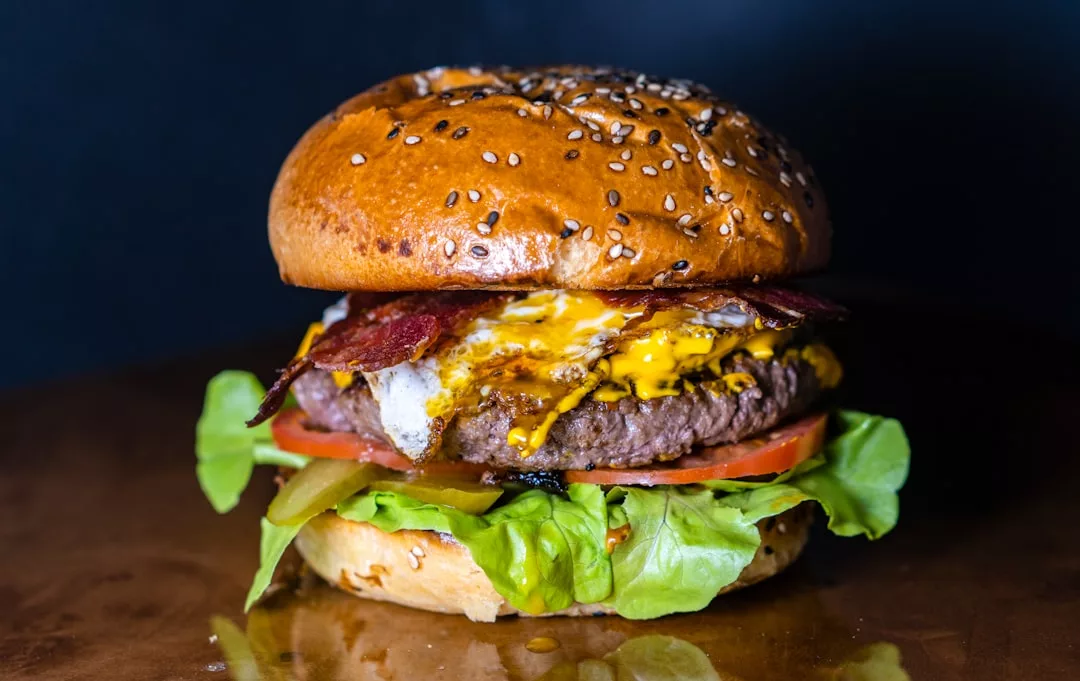 Nahaufnahme eines Cheeseburgers mit Sesambrötchen, Salat, Tomaten, Gurken, geschmolzenem Käse, Speck und einem Rindfleisch-Patty – perfekt zum Genießen nach einem Tag voller Erkundungen der Aromen Mallorcas – vor einem dunklen Hintergrund.