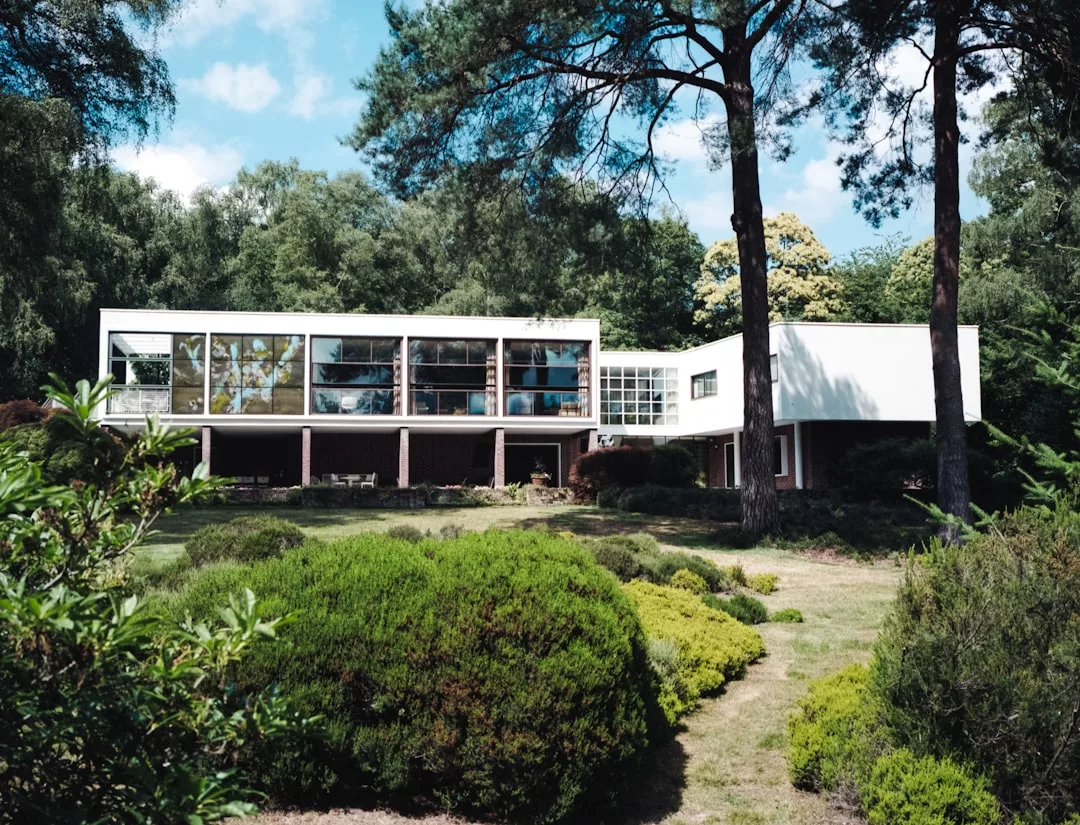 Modernes, weißes, zweistöckiges Haus mit großen Glasfenstern, auf Säulen stehend, umgeben von Bäumen und einem angelegten Garten – spiegelt den stilvollen Charme Mallorcas wider.