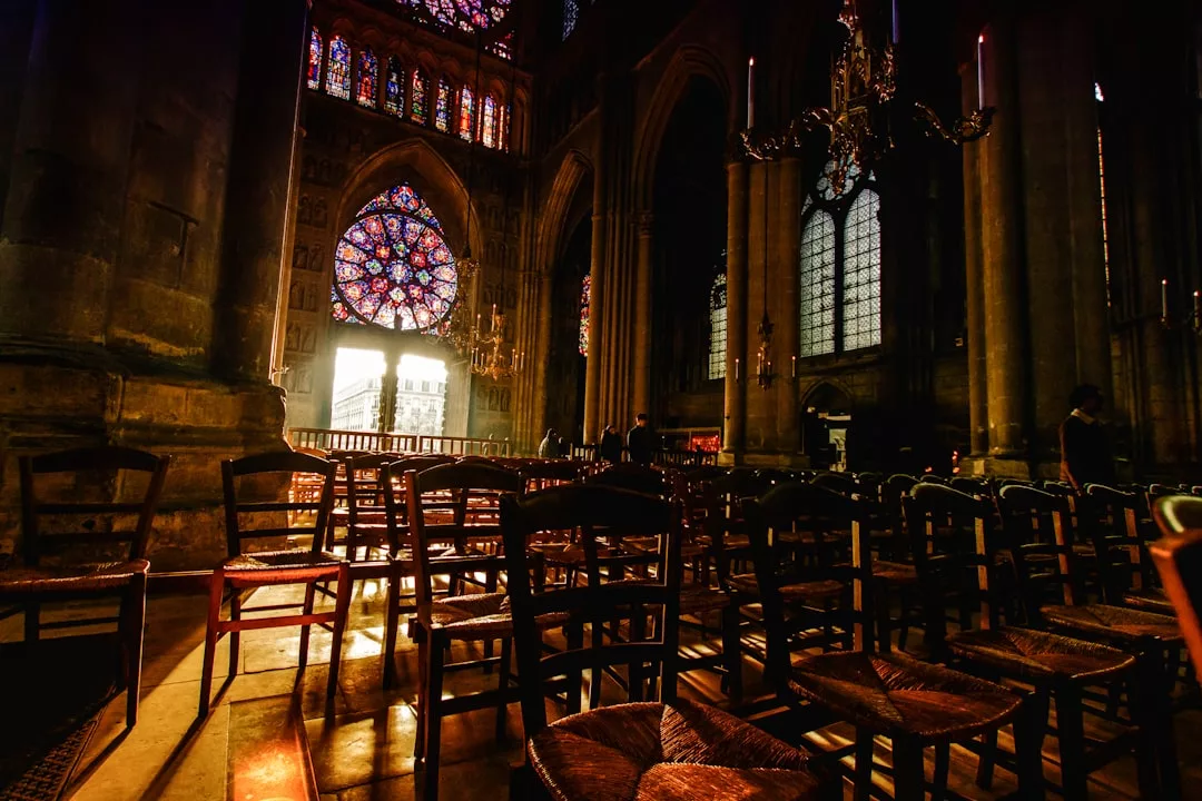 Reihen von Holzstühlen in einer schwach beleuchteten Kathedrale auf Mallorca mit Buntglasfenstern und Sonnenlicht, das durch den Eingang strömt.