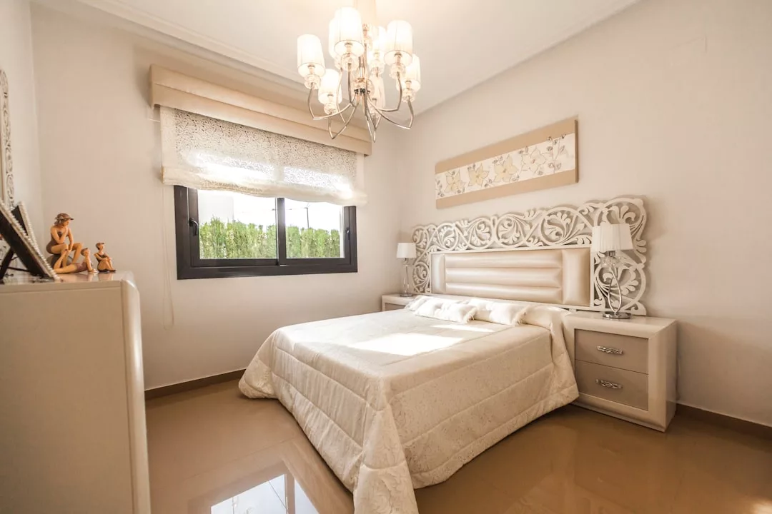 Ein helles Schlafzimmer im mallorquinischen Stil mit einem großen Bett, einem verzierten Kopfteil, zwei Nachttischen mit Lampen, einem Kronleuchter und einem mit transparenten Vorhängen geschmückten Fenster.
