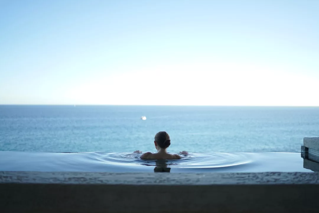 Eine Person mit hochgestecktem Haar entspannt sich in einem Infinity-Pool und blickt auf das ruhige Meer unter Mallorcas klarem blauen Himmel.