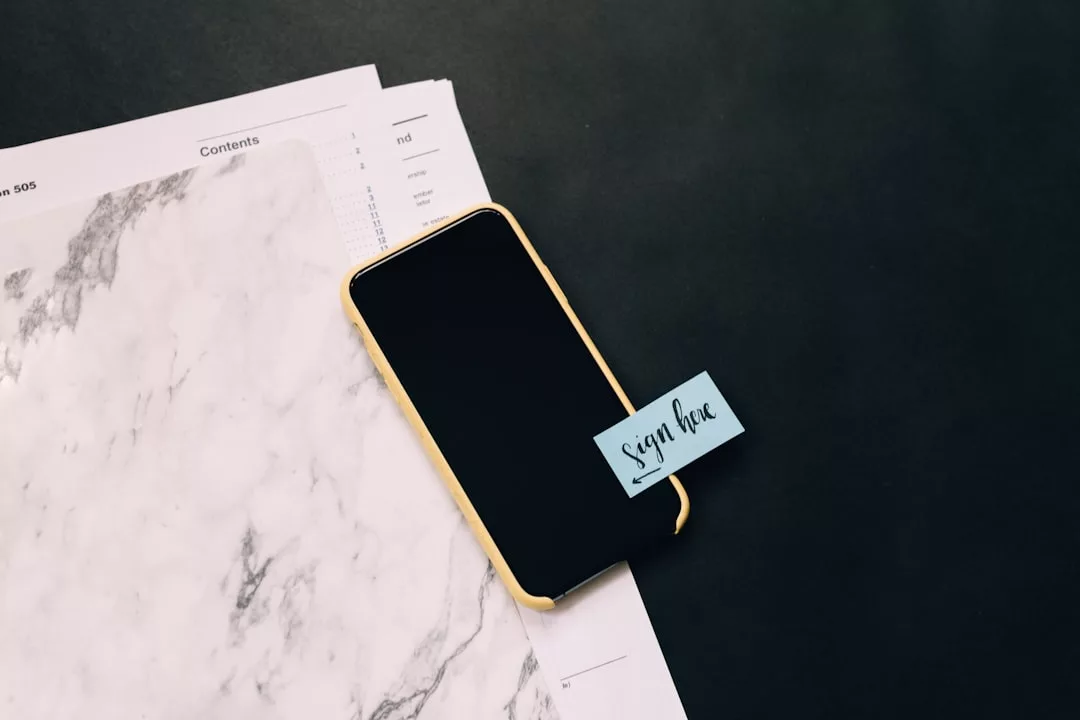 Ein Smartphone mit einer gelben Hülle liegt auf Papier mit Marmormuster, an dem ein blauer Haftzettel mit der Aufschrift „Hier unterschreiben“ klebt – eine Erinnerung an die lebendige Atmosphäre Mallorcas.