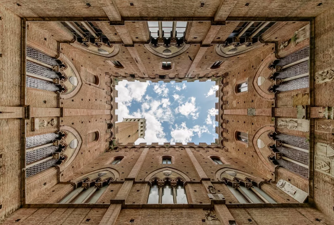 Blick nach oben auf einen rechteckigen Innenhof auf Mallorca, eingerahmt von hohen Backsteinmauern mit Bogenfenstern, Säulen und einem Blick auf blauen Himmel und Wolken darüber.