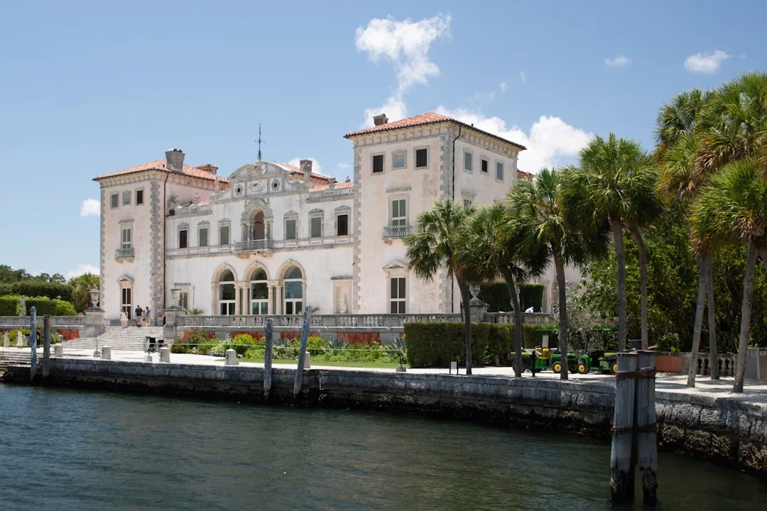 Ein großes, historisches Herrenhaus mit Bogenfenstern steht am Wasser auf Mallorca, umgeben von Palmen und einem Steinweg unter einem klaren blauen Himmel.