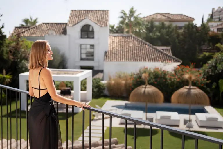 Eine Frau in einem schwarzen Kleid steht auf einem Balkon mit Blick auf einen angelegten Hinterhof auf Mallorca mit Pool, Liegestühlen und einer weißen Villa.
