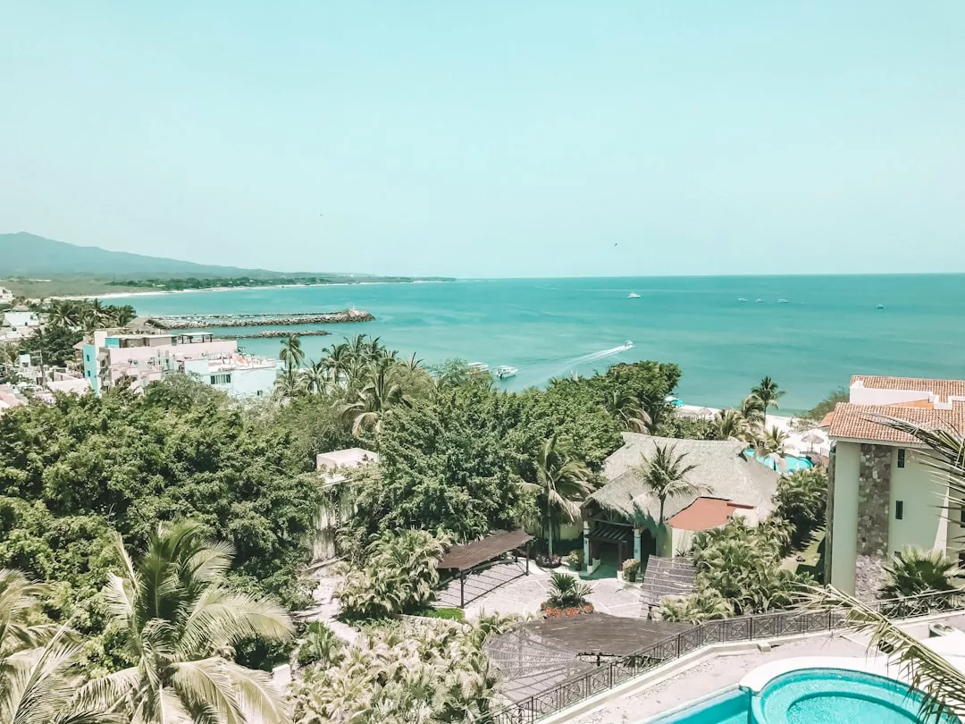 Luftaufnahme einer Küstenstadt auf Mallorca mit üppigen grünen Bäumen, Gebäuden, einem Swimmingpool im Vordergrund und dem Meer, das sich unter einem klaren Himmel bis zum Horizont erstreckt.