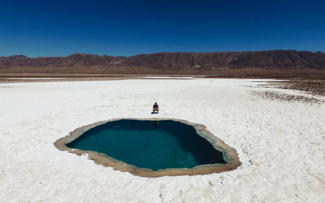 Eine Person sitzt am Rand eines kleinen, blauen Wasserbeckens, das von weißen Salzebenen umgeben ist und an die ruhigen Landschaften Mallorcas erinnert. Im Hintergrund sind unter einem klaren blauen Himmel Berge zu sehen.