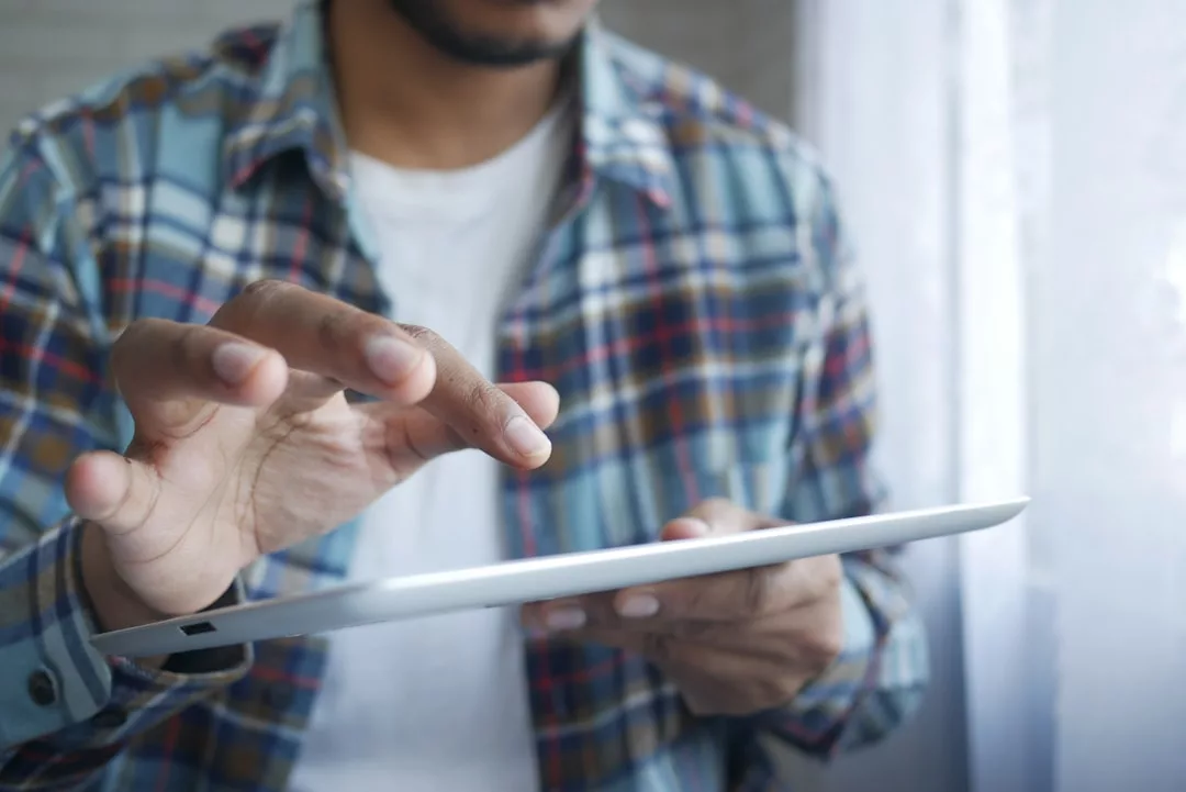 Eine Person in einem karierten Hemd verwendet ein Tablet-Gerät in der Nähe eines Fensters. Ihr Oberkörper und ihre Hände sind sichtbar. Vielleicht plant sie gerade ihren nächsten Urlaub auf Mallorca.