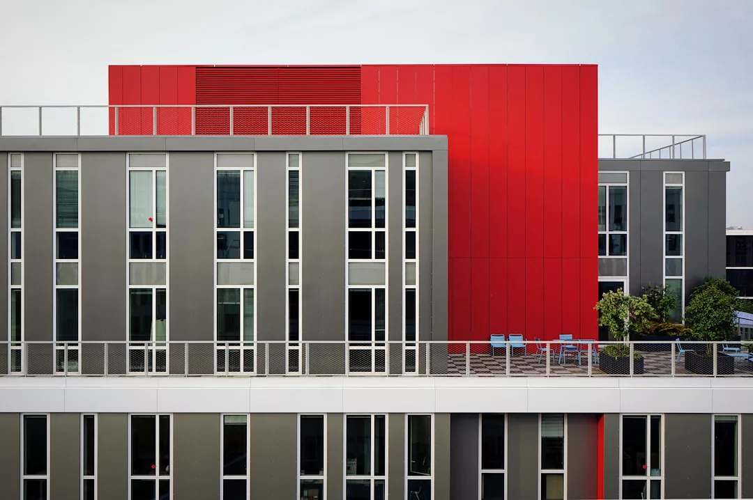 Modernes Apartmentgebäude auf Mallorca mit grauen und roten Außenwänden, großen vertikalen Fenstern und einer Dachterrasse mit Sitzgelegenheiten im Freien und üppigen Pflanzen.