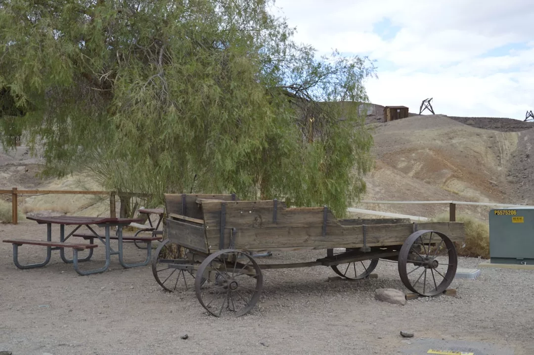 Ein alter Holzwagen mit Metallrädern steht auf Kies neben einem Picknicktisch und einem Baum und erinnert an die rustikale Landschaft Mallorcas, mit Hügeln und einem Verteilerkasten im Hintergrund.
