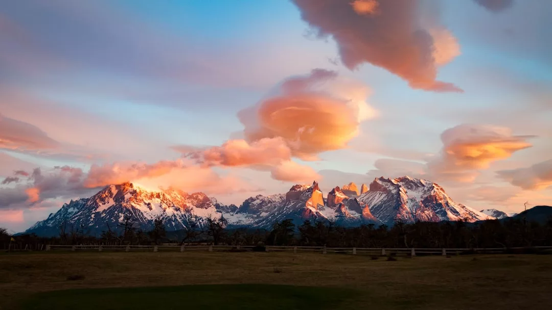 Schneebedeckte Bergkette bei Sonnenuntergang mit dramatischen, in Orange- und Rosatönen leuchtenden Wolken über einer Grasebene, die an die ruhigen Landschaften Mallorcas erinnert.