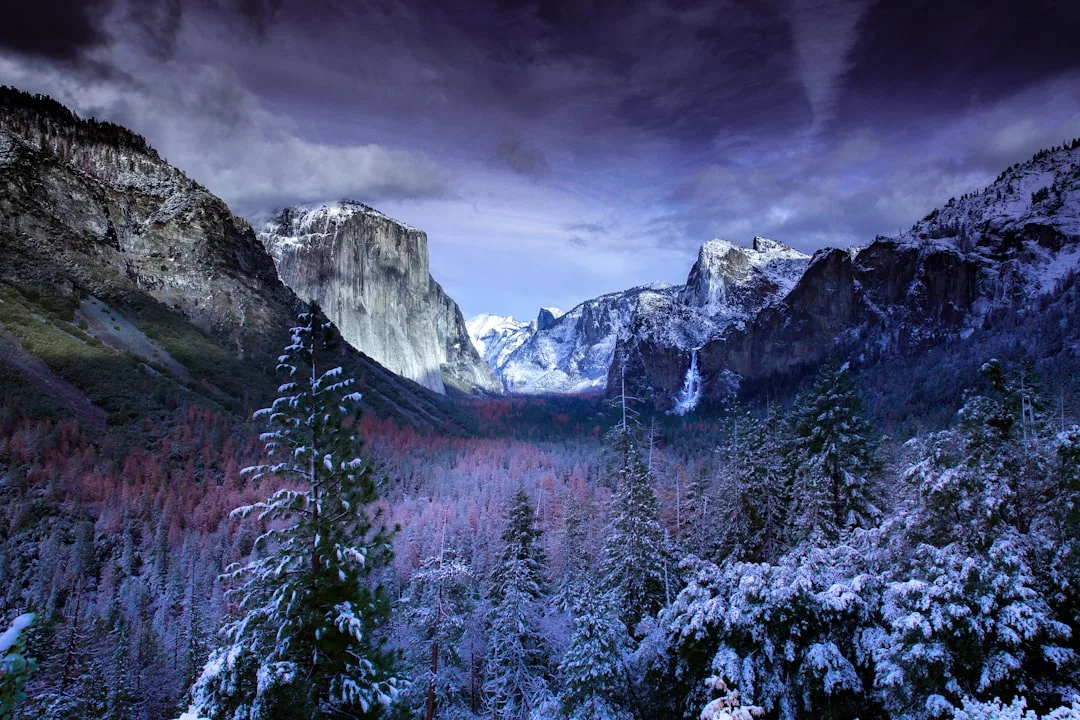 Schneebedeckte Bäume und Berge unter einem dramatischen, bewölkten Himmel füllen ein großes Tal, mit schroffen Klippen, die an die Landschaft Mallorcas erinnern, und fernen Gipfeln, die sich im Hintergrund erheben.