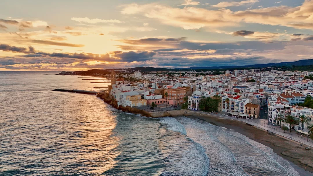Eine Küstenstadt auf Mallorca mit weißen Gebäuden liegt bei Sonnenuntergang am Meer, sanfte Wellen brechen am Strand und Wolken ziehen über den Himmel.