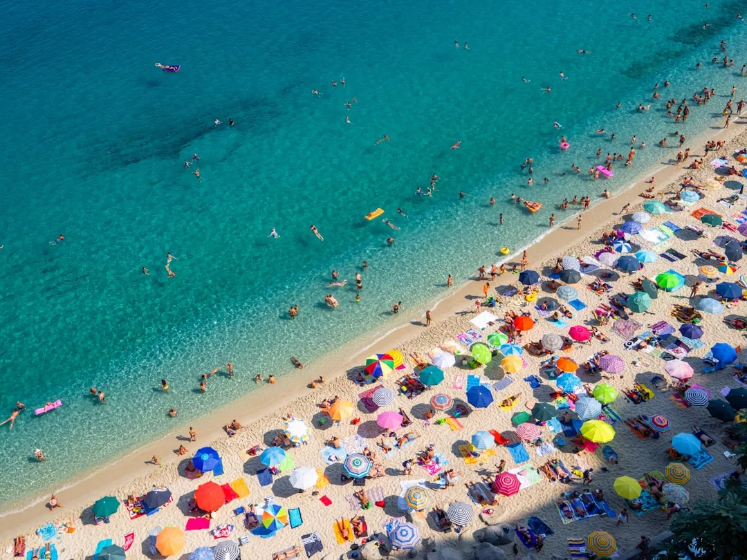 Eine überfüllte Strandszene auf Mallorca mit vielen bunten Sonnenschirmen im Sand und Menschen, die im klaren, blauen Wasser schwimmen und entspannen.